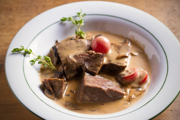 Braised beef cheeks in cream sauce with mushrooms and tomatoes in white bowl on wooden table