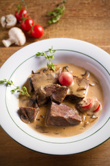 Braised beef cheeks in cream sauce with mushrooms and tomatoes in white bowl on wooden table