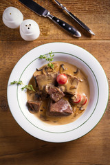 Braised beef cheeks in cream sauce with mushrooms and tomatoes in white bowl on wooden table. Overhead
