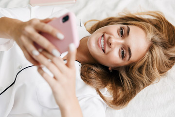 Fototapeta premium Image of beautiful girl holding cellphone while lying in bed at home