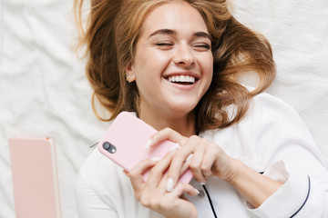 Image of joyful girl holding cellphone while lying in bed at home