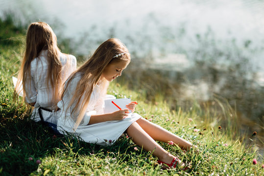 two little girls with pencil draws in notebook while sitting on the grass in the park, child writes in notebook