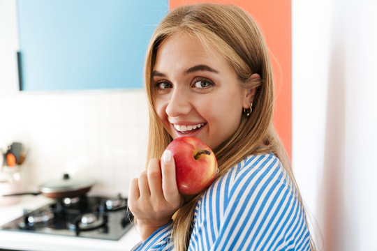 Image of joyous young girl smiling and eating apple in kitchen