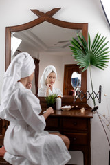 A girl with a towel on her head and in a white coat poses while sitting at a brown dressing table on which stands a white bottle of a mirror. The concept of care and health.