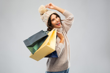 christmas, seasonal sale and consumerism concept - happy smiling young woman in knitted winter hat and sweater with shopping bags over grey background