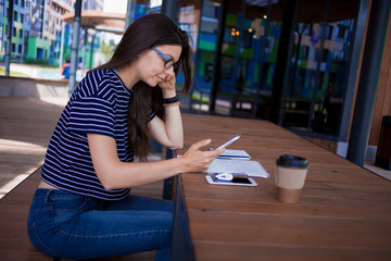 A serious brunette girl, freelancer, with large wrist watch, works at a wooden table on tablet,...