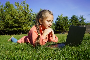 A girl of seven years old works on a laptop on the green grass. Distance learning. Girl with a laptop on a background of pines. Girl with a computer on the street.