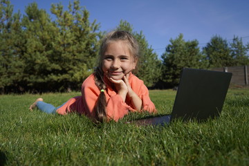 A girl of seven years old works on a laptop on the green grass. Distance learning. Girl with a...