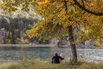 woman and dog sitting under a tree with yellow and green leaves looking at the water of the mountain lake in autumn