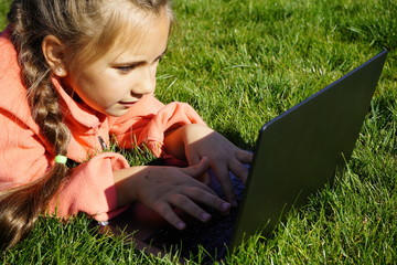 A girl of seven years old works on a laptop on the green grass. Distance learning. Girl with a...