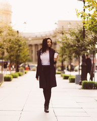 Beautiful serious smart brunette girl holding cup of coffee in hands goes walking down street of St. Petersburg in city center. Charming thoughtful woman with long dark hair wanders alone