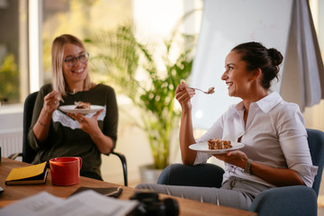 Two smiling women in the office eating cake - obrazy, fototapety, plakaty