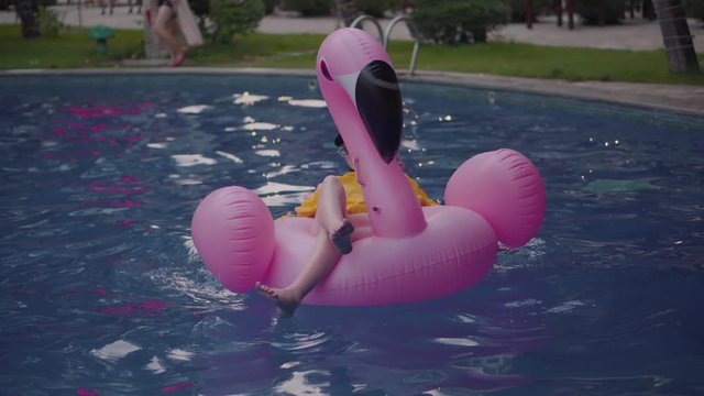 A young girl swims in the afternoon in the pool. Fashionable woman in a cap is rowing on a pink inflatable flamingo. Mattress, flamingo, girl in an open air hotel.