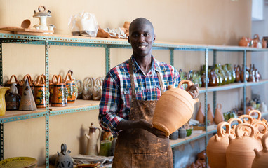 Afro-american artisan in apron having ceramics in store warehouse