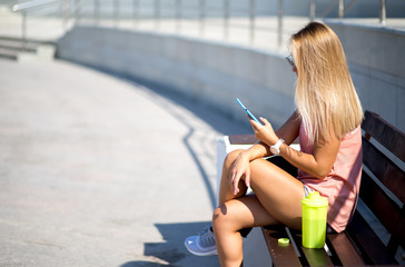 Sportswoman looking at smart phone, outdoors. Fitness female resting after run in the city.