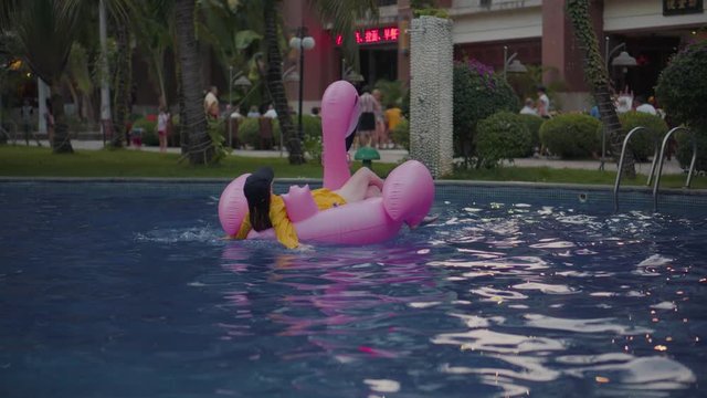 A young girl swims in the afternoon in the pool. Fashionable woman in a cap is rowing on a pink inflatable flamingo. Mattress, flamingo, girl in an open air hotel.