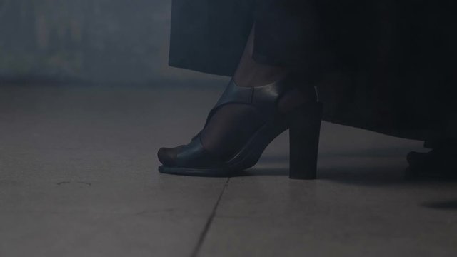 A woman in a black cloak, pantyhose and open sandals with thick heels is slowly walking along the tile in the room. The room is twilight. Close-up of legs. A little smoke spills across the floor.