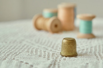 Vintage brass thimble close-up.  It is on an old handmade knitted tablecloth.  In the background vintage wooden bobbins with sewing threads