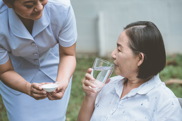Asian nurse helping sick elderly woman to drink water with medical pill.
