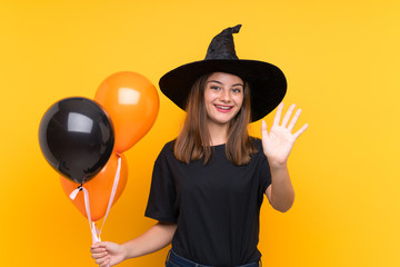 Young witch holding black and orange air balloons for halloween parties saluting with hand with happy expression