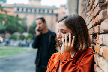 Girl talking with the mobile with serious expression and with a boy in the background talking with the mobile with a brick wall next to it.