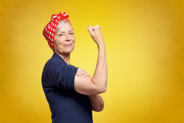 Senior rosie riveter concept: self-confident elderly woman with clenched fist rolling up her sleeve, copy space