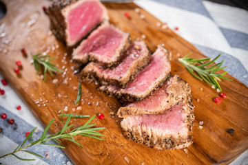 Full steak with black pepper cutted with a knife and fork on a chopping board with many rosemary leaves and spices, medium rare, red meat, gray-white napkin background. Good food make to good health