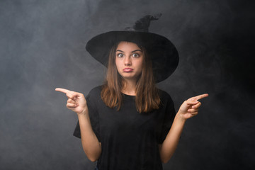 Girl with witch costume for halloween parties over isolated dark background pointing to the laterals having doubts