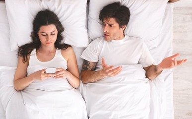 Angry man talking to his wife browsing cellphone and ignoring him in bed