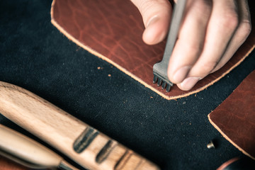 a tanner punching holes on a brown leather workpiece on a desktop. The tanner holds in his hand a...