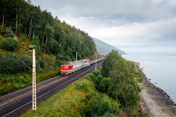 Freight train on the railroad of Trans-Siberian Railway on the shore of Baikal Lake with green forest trees and cloudy sky. East Siberian Railway in Buryatia, Siberia, Russia