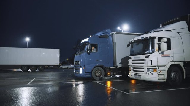 Blue Semi-Truck with Cargo Trailer Drives Into Overnight Parking Space where Other Trucks are Standing. Drivers Resting at Night on the Overnight Parking Lot