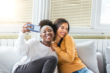 Laughing woman friends hugging each other on sofa while taking selfie photo on smart phone. Lovable...