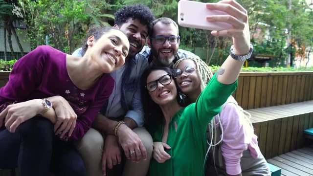Woman with eyeglasses suggests taking a selfie with friends. Smiles, grimaces and fun. Diverse men and women friends having a break for a selfie photograph all together in the outdoor. Time of fun, bo