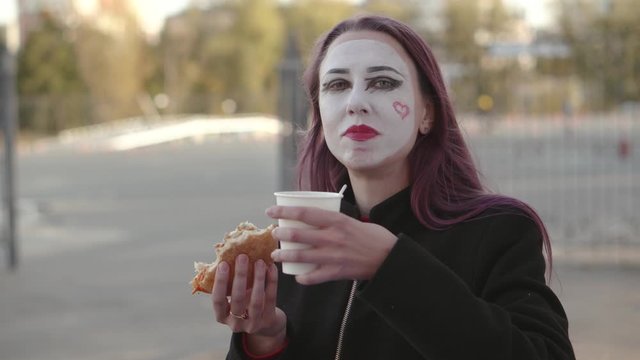 Mime girl in a black coat eating a hamburger winking at the camera and drinking coffee.