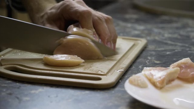 A man slices a chicken fillet with a large knife on a chopping Board