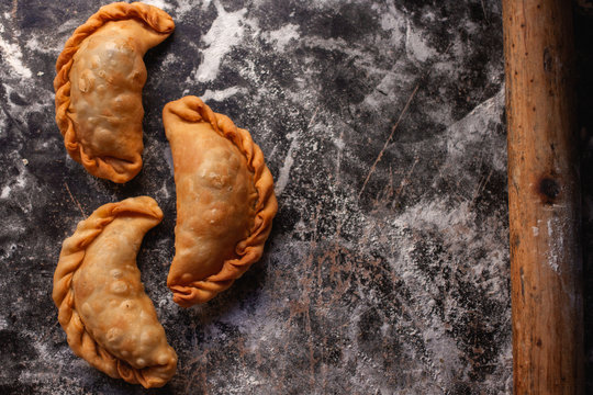 tres empanadas fritas junto a un palo de amasar sobre una mesa con harina