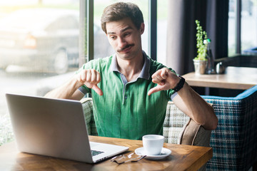 Dislike! Young dissatisfied businessman in green t-shirt sitting, working and looking at laptop...