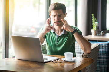 Young worry hopeful businessman in green t-shirt sitting with closed eyes and crossed fingers and hope to win. business and freelancing concept. indoor shot near big window at daytime.