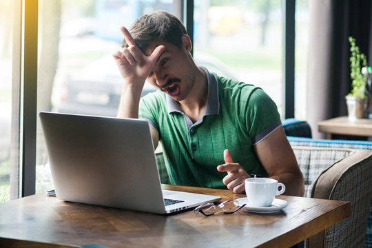 I am loser. Young angy businessman in green t-shirt sitting and working on laptop with loser gesture on his forehead and screaming. business and freelancing concept. indoor shot near window at daytime