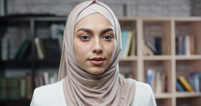 Female muslim student in library, holding books and looking at camera, girl wearing hijab spending time studying - modern islam, student lifestyle concept close up 4k