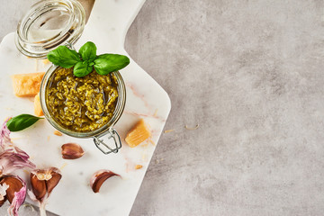 Pesto sauce or pesto genovese in a glass jar with pine nuts, parmesan, basil, oil and garlic on white marble cutting board over grey stone background. Top view. Copy space.