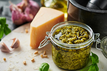 Pesto sauce or pesto genovese in a glass jar with pine nuts, parmesan, basil, oil and garlic on white marble cutting board. Copy space.