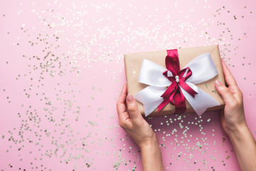Gift or present box with a big bow in the hands of a woman on a pink table. Flatlay composition for Christmas birthday, mother day or wedding.
