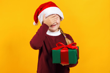 Funny little girl holds a box with a gift and closed her eyes with her hand. A child in a santa hat and a dark red sweater on a yellow background