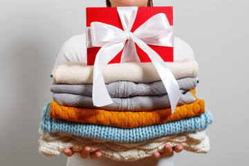 A woman holds a stack of warm knitted sweaters and a box with a gift on a gray wall background