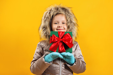 The child holds a box with a gift in his hand. Portrait of a happy cute little girl in a down jacket on a yellow background
