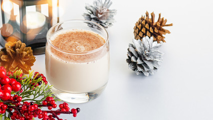 Traditional Christmas cocktail Eggnog with eggs, alcohol, grated nutmeg and cinnamon closeup. Sweet traditional drink on grey table with beige and red decorations and pine cones
