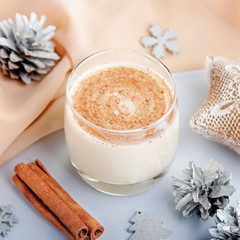 Traditional Christmas cocktail Eggnog with eggs, alcohol, grated nutmeg and cinnamon closeup. Sweet traditional drink on grey table with beige decorations, burning lanterns and pine cones