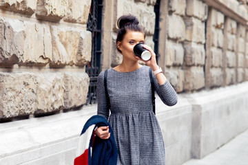 Pretty girl student is drinking from paper cup near stone wall of university. Female student drinks coffee on the city street near stone wall.